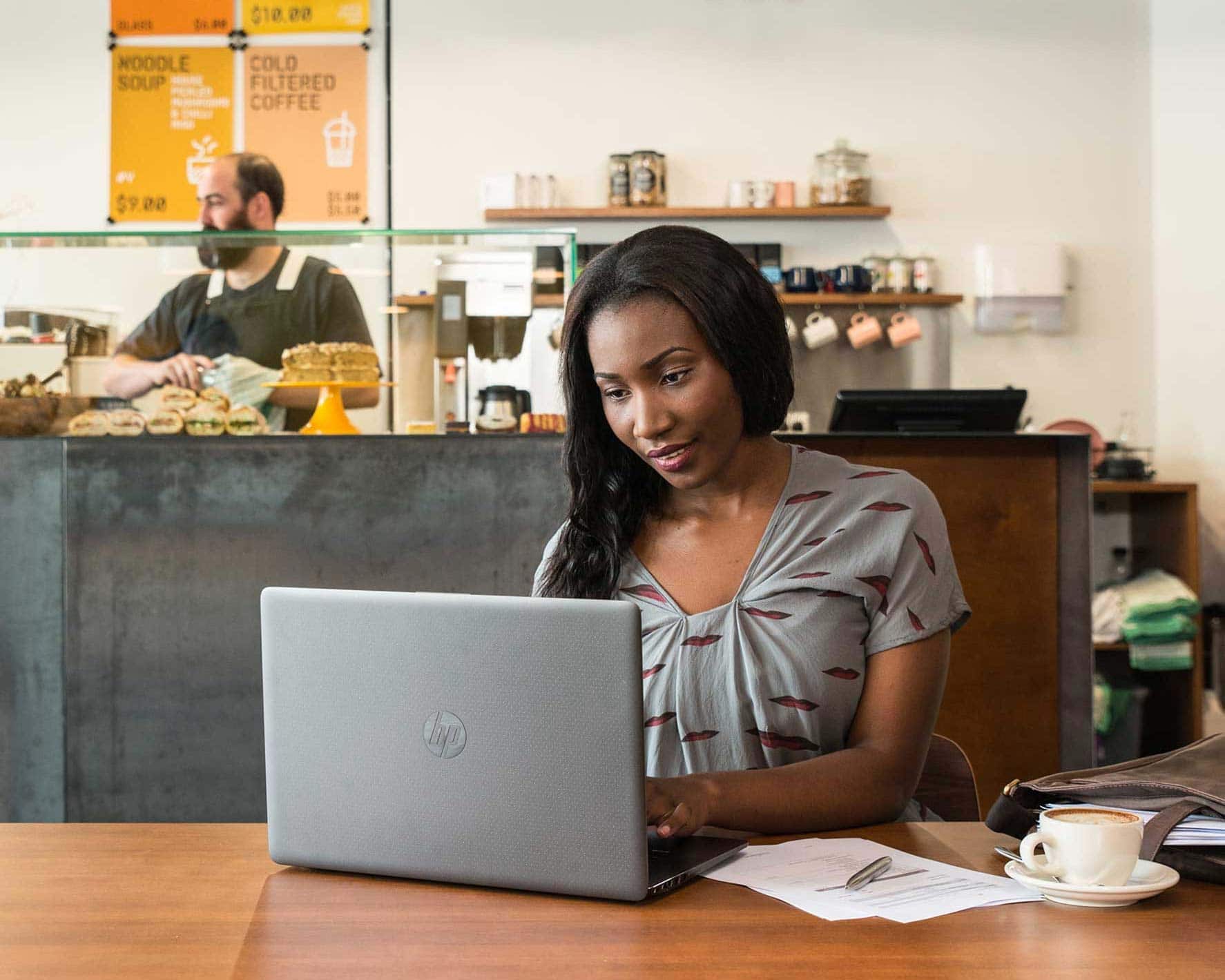 A woman sits in a coffee shop on her laptop, with a coffee cup, and a stack of papers next to her.